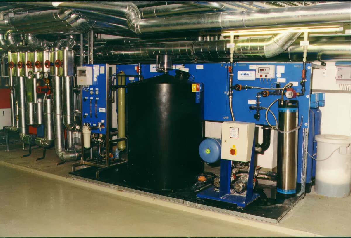 Wasseraufbereitungsanlage mit blauen Schaltschränken, schwarzem Vorlagebehälter, Filterkerzen und Enthärtungssäulen unter gedämmten Rohrleitungen